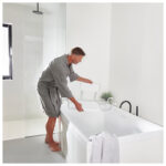 Man i grå morgonrock placerar vit badkarssits med ryggstöd på modernt badkar