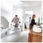 Modernt badrum med badkarssits monterad på vitt badkar och kvinna vid handfat