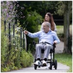 eFOLDi Navigator – i användning med vårdarkontroll utomhus Man i elrullstol med assistent på promenadväg bland blommor